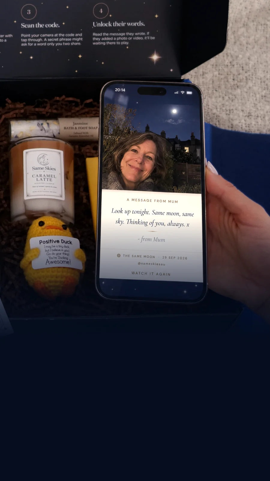 A hand holding a phone showing a Same Skies message from Mum with her selfie under the full moon, over an open Same Skies gift box with a candle, a crocheted Positive Duck and chocolate