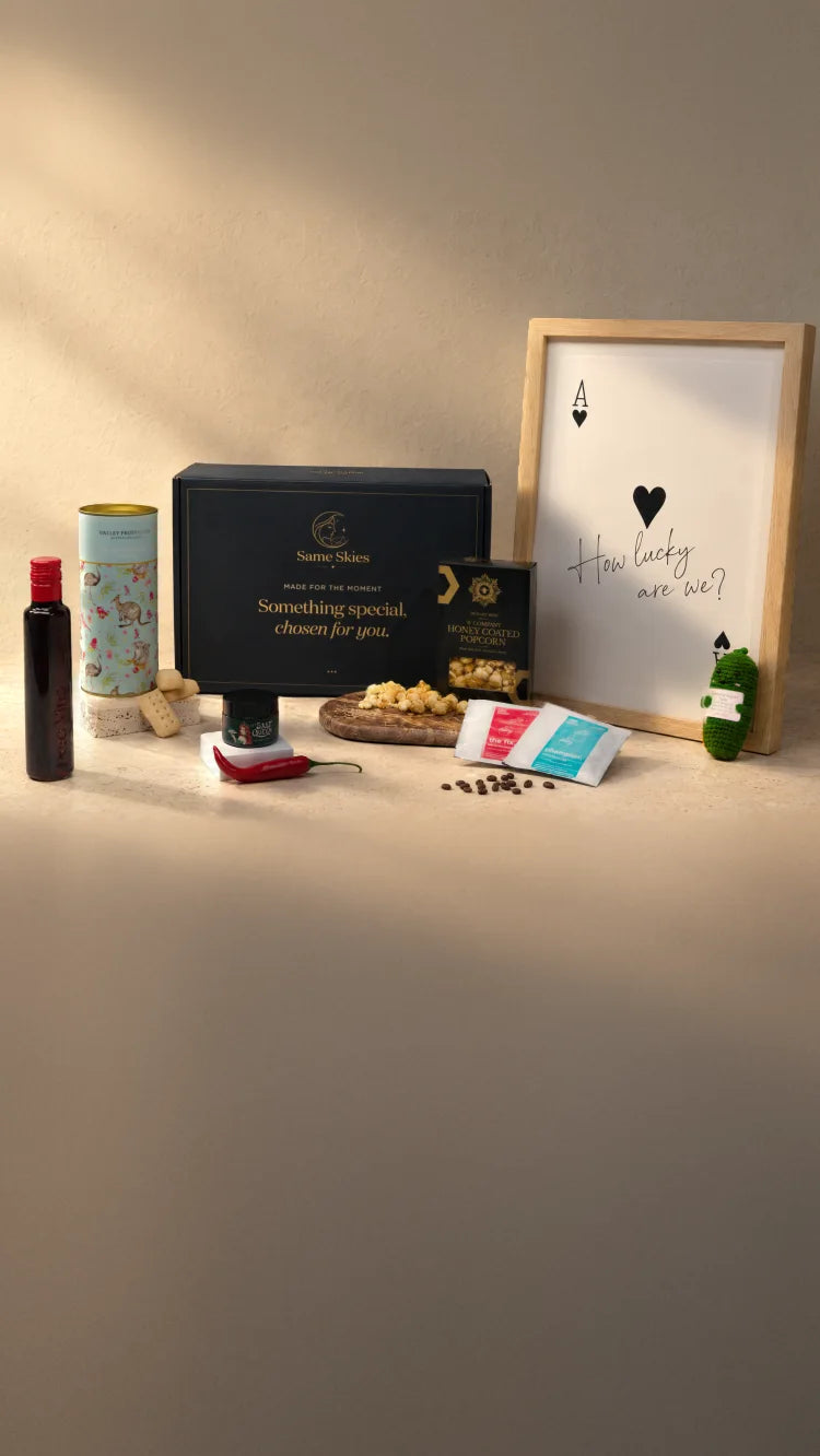 A Same Skies gift box with everything inside on warm plaster in late sun - a Poco Vino piccolo, Australian Shortbread, Jalapeño & Lime Salt, Honey-Glazed Popcorn, a Pour-Over Coffee Duo, an A3 Matte Art Print (How Lucky Are We?) and the navy Same Skies box