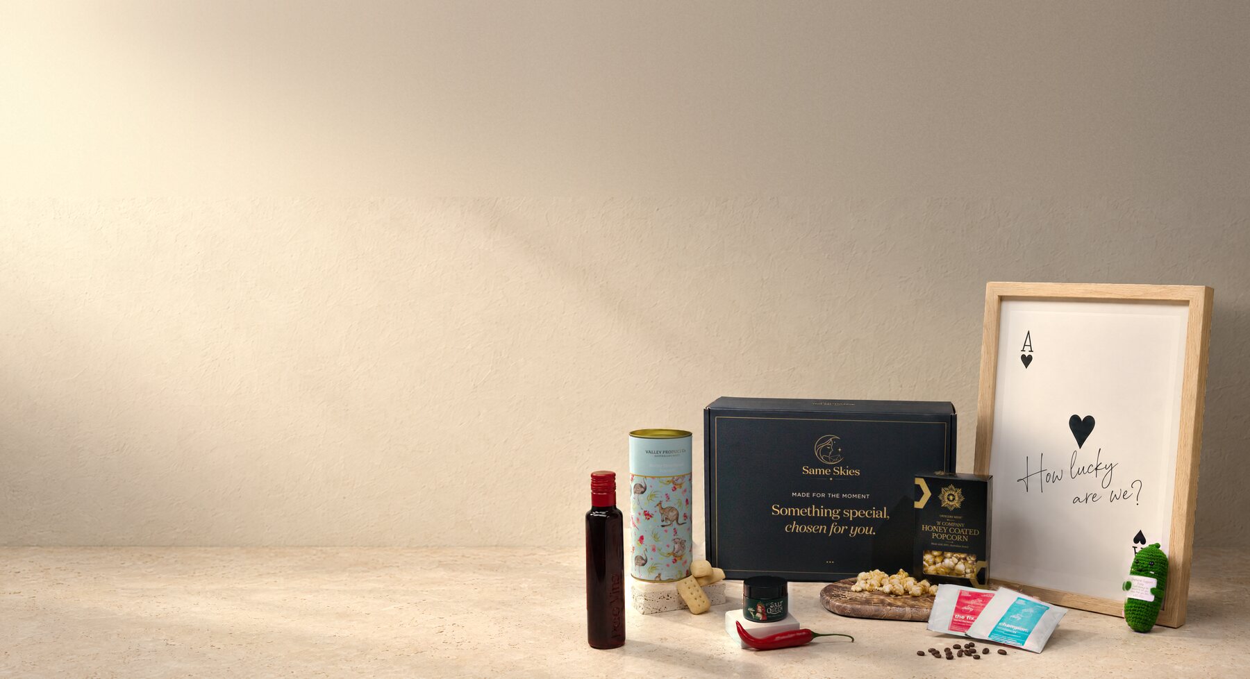Everything inside a Same Skies gift box on a warm ivory plaster and limestone backdrop - a Poco Vino piccolo, Australian Shortbread, Jalapeño & Lime Salt, Honey-Glazed Popcorn, a Pour-Over Coffee Duo, an A3 Matte Art Print (How Lucky Are We?) and the navy Same Skies box