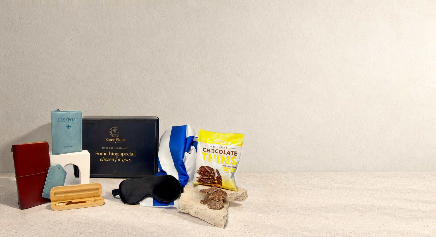 The Traveller gift box on warm plaster and limestone in morning light - a teal Passport Wallet & Tag Set, a Hardcover Travel Journal, a Hardwood Pen & Bamboo Case, a black Mulberry Silk Sleep Mask, a blue Cabana Beach Towel, Honeycomb Chocolate Thins and the navy Same Skies box