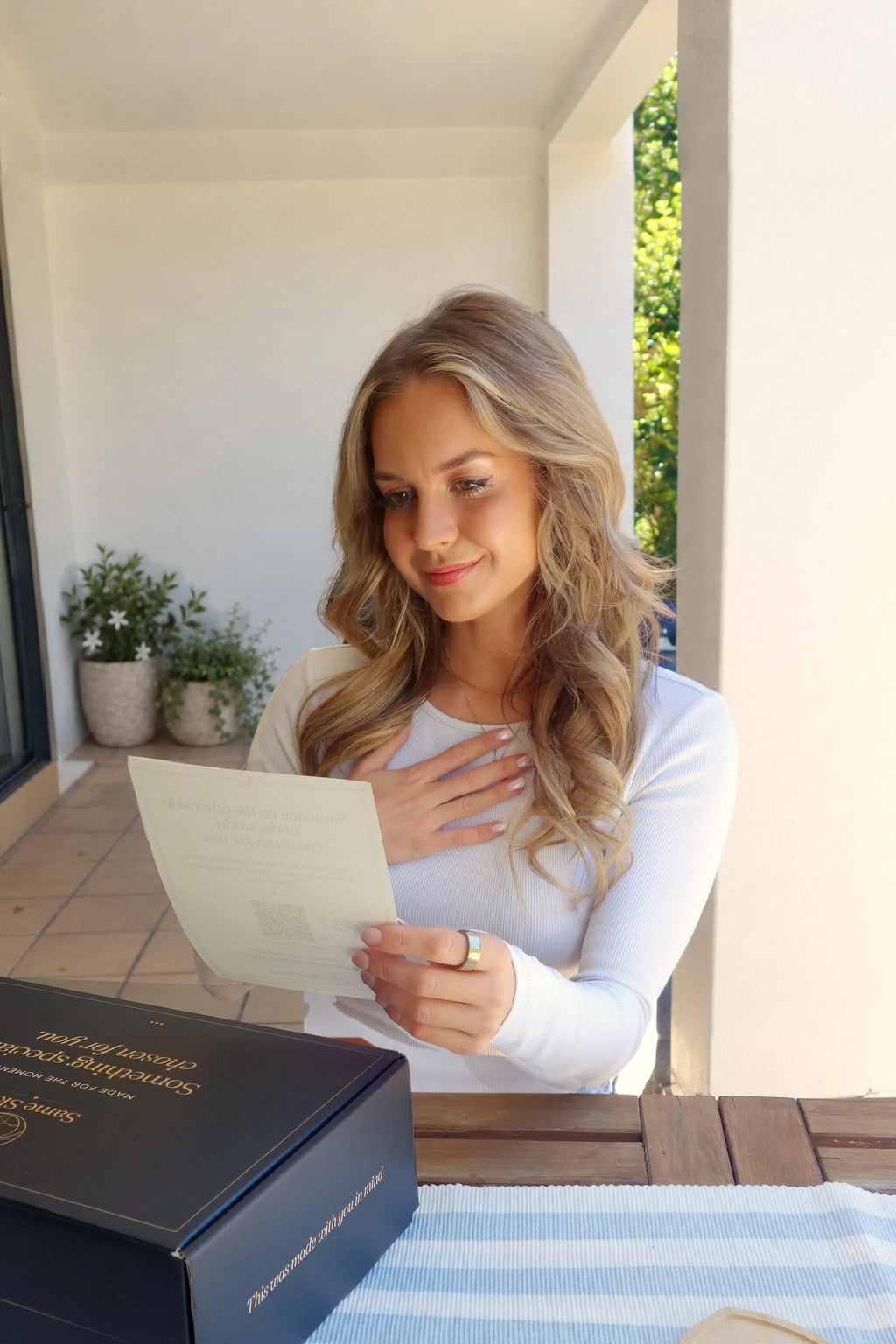 A woman with her hand on her heart, smiling as she reads the Same Skies letter card beside a navy Same Skies gift box