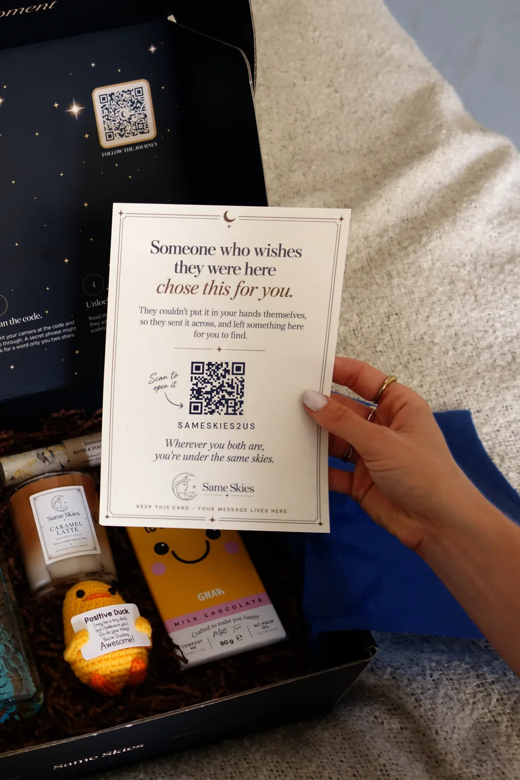 A hand holding a Same Skies letter card over an open navy gift box with Valley Produce shortbread, a Same Skies candle, a crocheted Positive Duck and GNAW milk chocolate