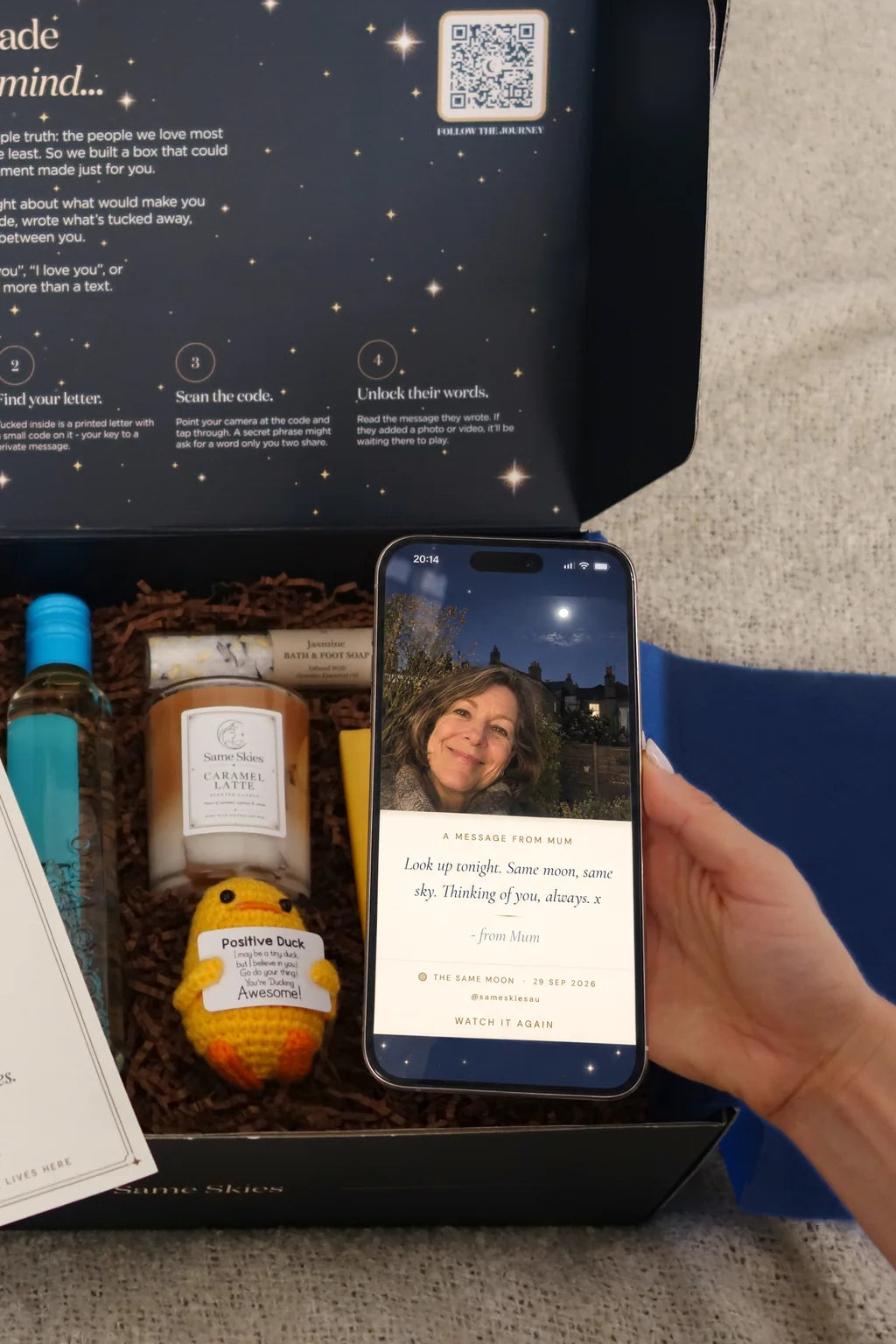 A hand holding an iPhone showing a Same Skies message from Mum with her selfie under the full moon, over an open gift box with a candle, a crocheted Positive Duck and chocolate